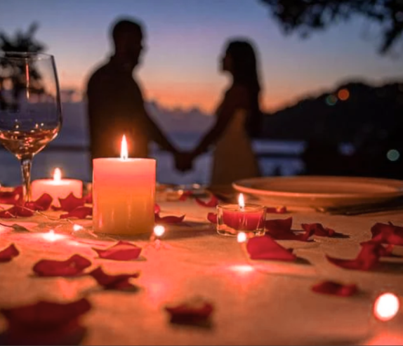 Tabel with white and pink candles, rose petals scattered about, a plate and wine glass with a couple holding hands in the background as the sun is setting with hills of trees. 