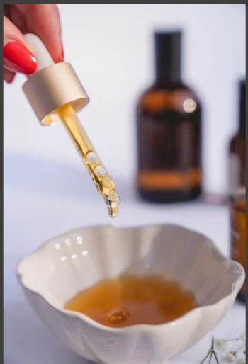 A dropper with oil being dropped into a white bowl and oil bottles in the background. 