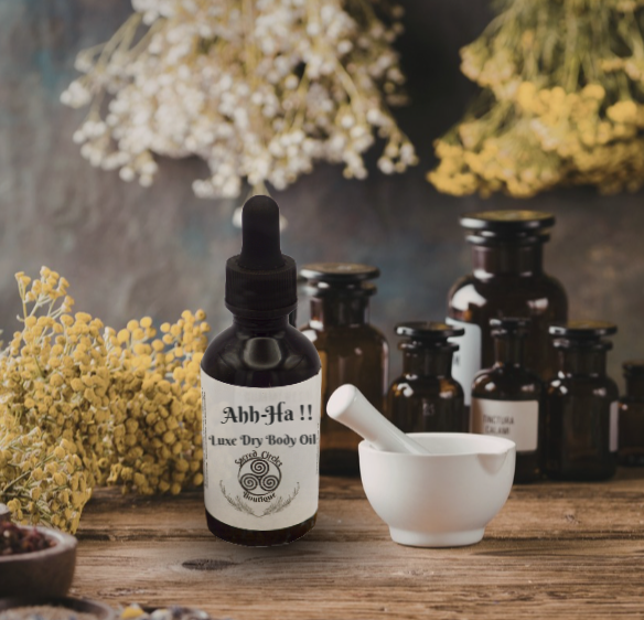 Bottle of Abh-Ha Luxe Dry Body Oil with a dropper on a wooden surface with flowers and other bottles in the background.