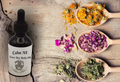 Bottle of Calm AF Luxe Dry Body Oil with dried flowers and herbs on a wooden surface
