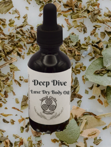 Bottle of 'Deep Dive Luxe Dry Body Oil' on a background of dried herbs