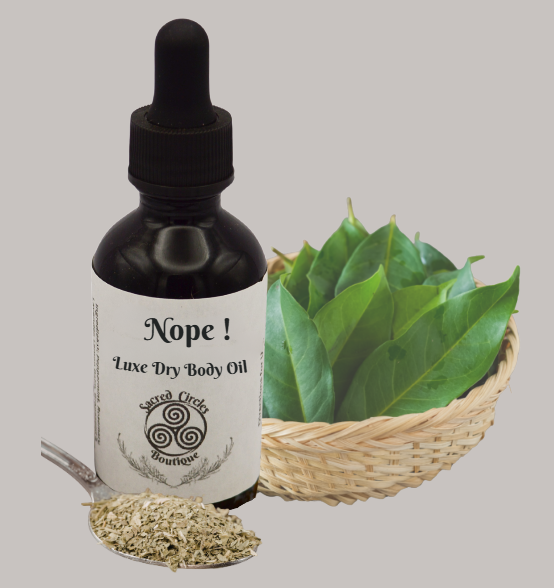 Bottle of 'Nope!' Luxe Dry Body Oil with a dropper, surrounded by green leaves and dried herbs on a gray background.