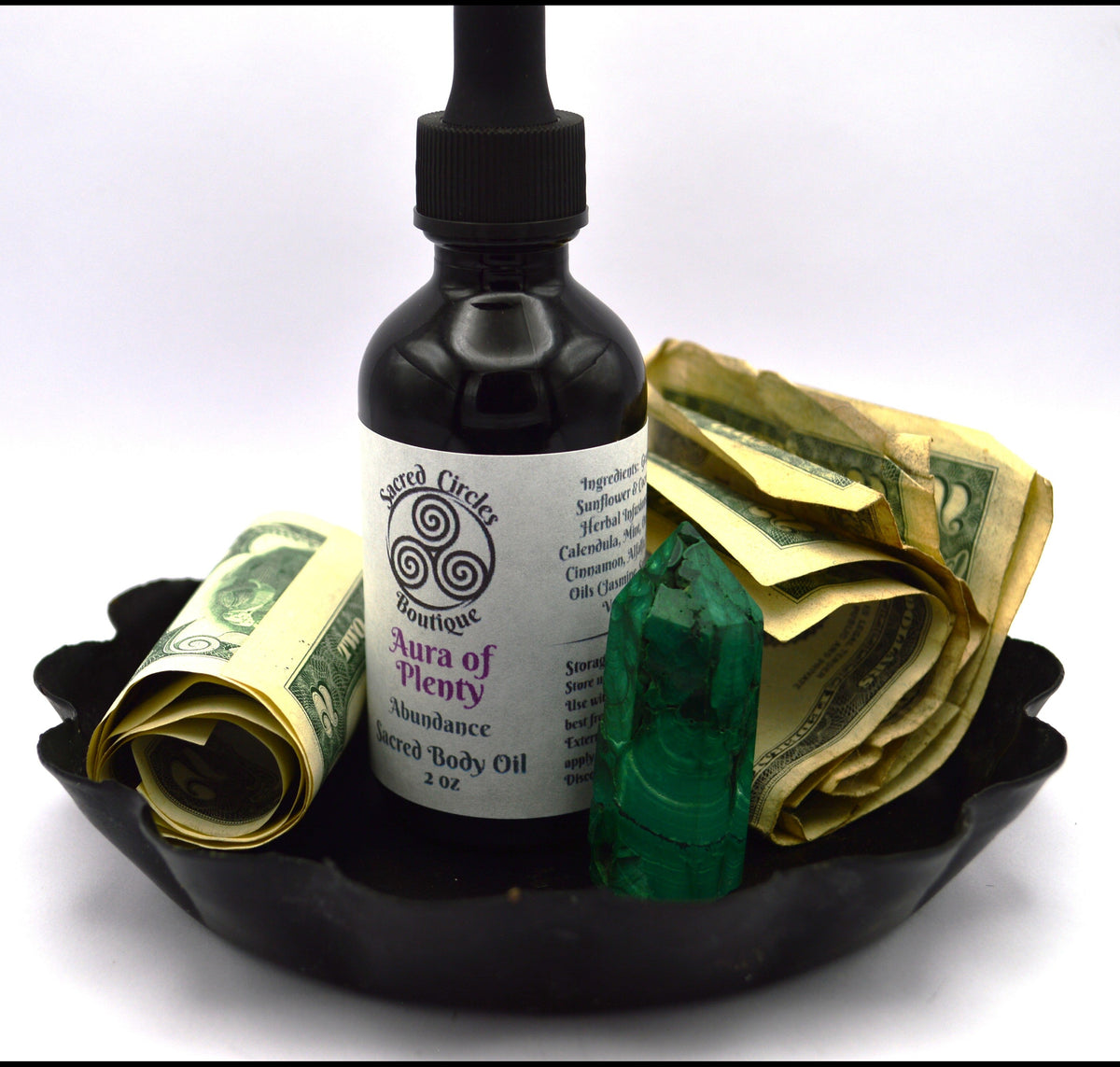 Black bottle of 'Aura of Plenty' body oil with rolled-up dollar bills on a white background